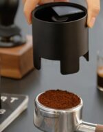 A hand lifts a black cylindrical coffee distribution tool above a portafilter basket filled with freshly ground coffee. The stainless steel portafilter rests on a countertop, with coffee-making accessories blurred in the background.