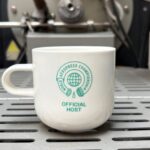 A white ceramic coffee mug displaying the teal "World AeroPress Championship – Official Host" logo sits on the cooling tray of a commercial coffee roasting machine. The industrial roaster is softly out of focus in the background, highlighting the commemorative design on the mug.