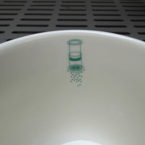 Close-up view of the inside rim of a white ceramic coffee mug featuring a small teal illustration of an AeroPress brewing coffee. The minimalist design sits just below the rim, with the slotted surface of a coffee roasting machine softly blurred in the background.