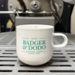 A white ceramic coffee mug with a teal "Badger & Dodo" logo sits on the cooling tray of a commercial coffee roasting machine. The industrial roaster is softly blurred in the background, emphasizing the branded mug in the foreground.