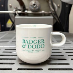 A white ceramic coffee mug with a teal "Badger & Dodo" logo sits on the cooling tray of a commercial coffee roasting machine. The industrial roaster is softly blurred in the background, emphasizing the branded mug in the foreground.