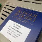 Navy blue Badger & Dodo coffee box with a white label reading 'Colombia, Jario Arana, Co Fermented Watermelon, Region: Armenia, Quindio, Farm: Santa Barbara, Varieties: Castillo, Cupping notes: Watermelon Candy, Key Lime, Banana, Potpourri, Cupping score: 88' with a QR code, sitting on a counter in a coffee roastery