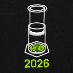 The image shows a close-up view of the embroidered design on a BlackT-shirt. The design features a white illustration of an AeroPress coffee maker, with a stylized coffee bed inside a hexagon at the bottom. Below the graphic, the year “2026” is stitched in bold white text.