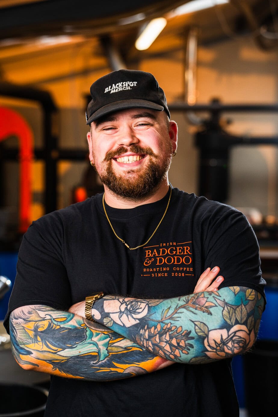 Portrait of a smiling bearded man with his eyes crinkled in a warm grin, standing with his arms crossed in front of coffee roasting equipment. He wears a black cap reading "Blackspot Merch", a gold chain, a gold watch, and a black t-shirt embroidered in orange with "Irish Badger & Dodo, Roasting Coffee, Since 2008". Both forearms are covered in colourful tattoos of feathers, roses, pine cones and leaves in blue, teal and orange tones. The background is softly blurred with warm lighting and industrial machinery.